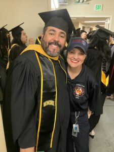 Hallway of multiple graduate students waiting for ceremony to start. Primary picture subjects - One male EDHS graduate student in black graduation robes and gold and black graduate stole (Thomas Rivalis) next to prior EDHS graduate student (Hilary Budzinski Betley) in blue fire company BDUs.