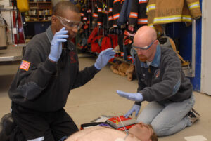 Two men with gloves doing CPR, hands raised as they use a defibrillator to check a simulated man having a cardiac arrest