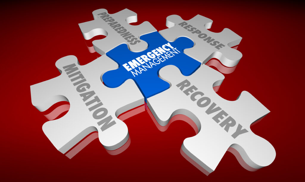5 jigsaw puzzle pieces together in an X shape with red background. Blue puzzle piece in center with words Emergency Managenment. Top left, white puzzle piece with word Preparedness. Top right, white puzzle piece with word Response. Bottom right, white puzzle piece with word Recovery. Bottom left, white puzzle piece with word Mitigation.